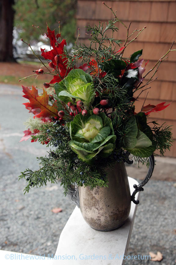a silver teapot of flowering cabbage, oak leaves, juniper, blueberry, and rose hips